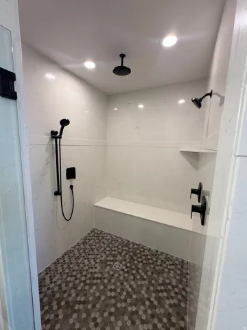 Spacious walk-in shower featuring a rain shower head, additional shower head with handheld sprayer, matte black fixtures, built-in bench, and hexagonal mosaic tile flooring