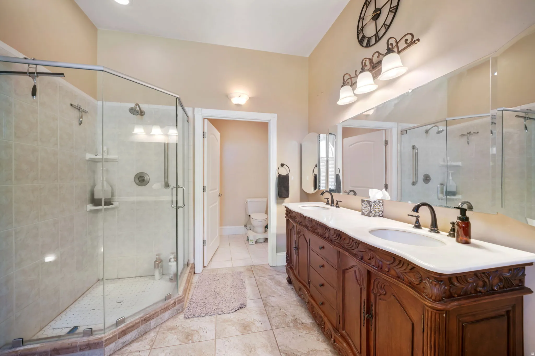 Spacious main primary bathroom featuring a dual vanity with carved wood cabinetry and white countertop, a large glass-enclosed shower with tiled walls, and neutral-toned floor tiling