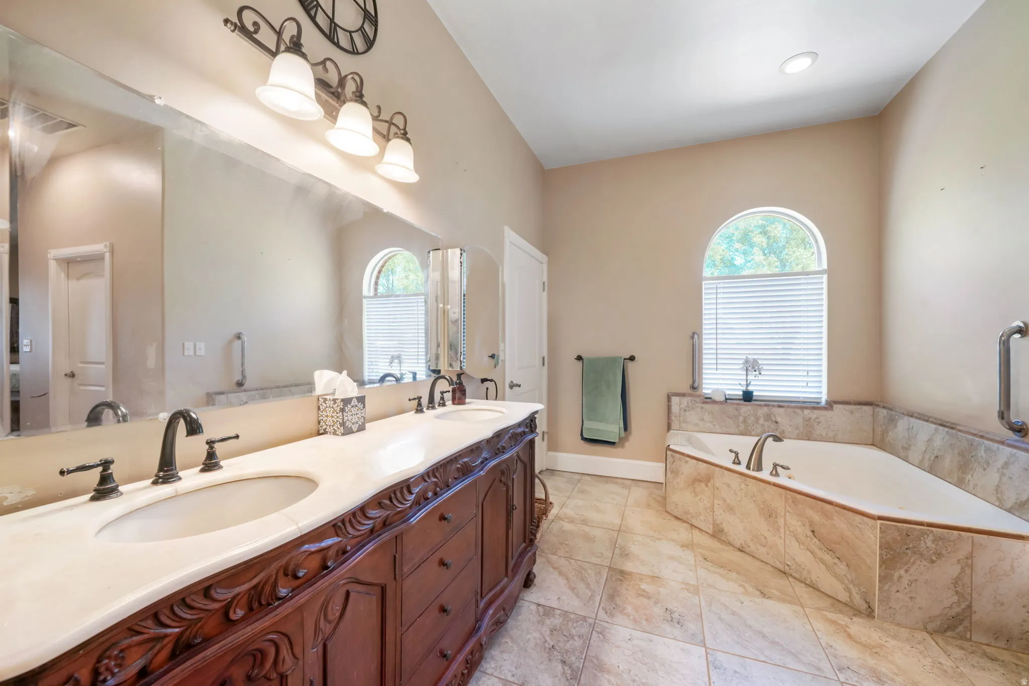 Spacious main primary bathroom featuring a carved wood-finish double vanity with dual sinks, a large mirror, a corner soaking tub with a tiled surround, and an arched window with blinds