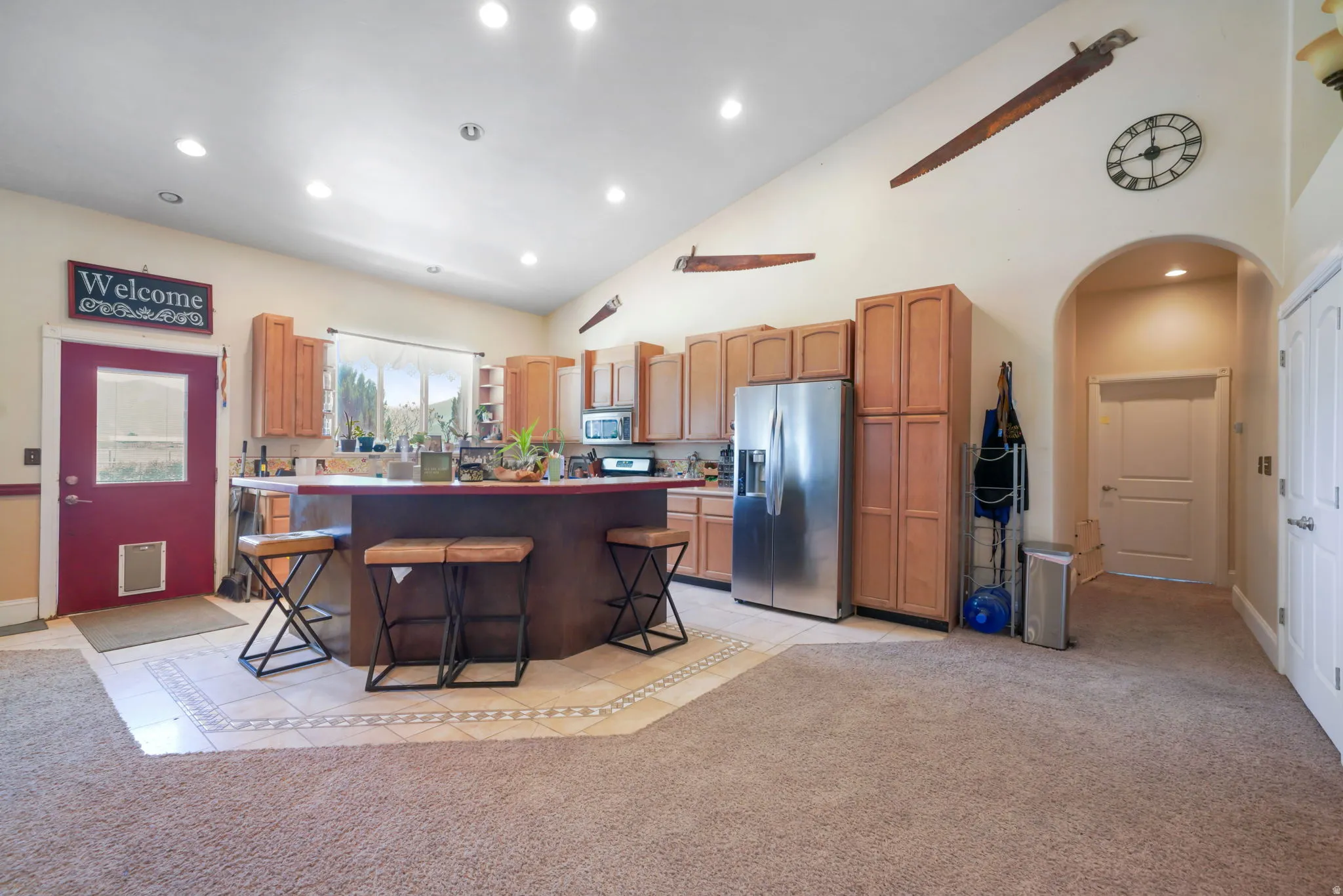 Expansive kitchen and living area featuring vaulted ceilings with recessed lighting, wood-finish cabinetry, a stainless steel refrigerator, and a kitchen island with seating