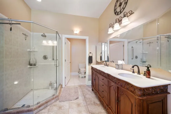 Spacious main primary bathroom featuring a dual vanity with carved wood cabinetry and white countertop, a large glass-enclosed shower with tiled walls, and neutral-toned floor tiling