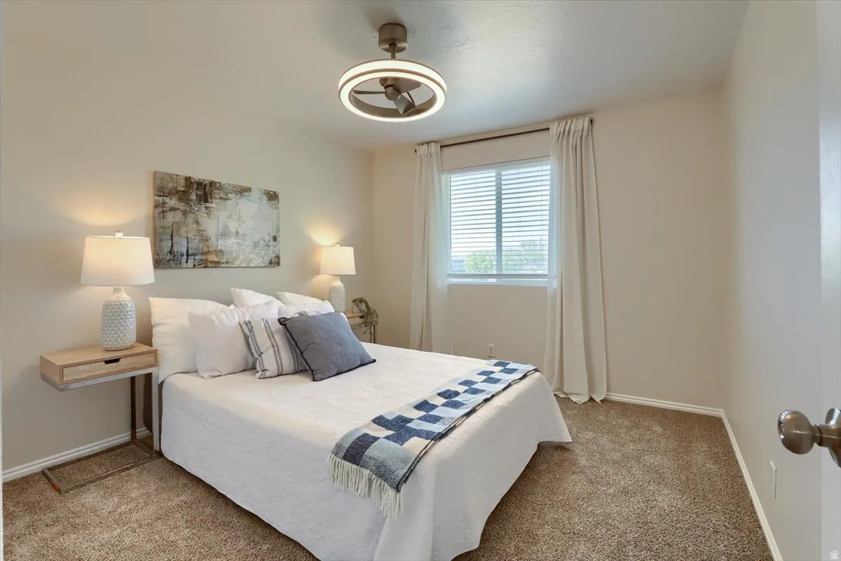 Carpeted bedroom featuring neutral wall tones, a modern ceiling fan with integrated lighting, a window with blinds and drapes, and white baseboards