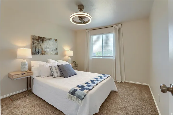 Carpeted bedroom featuring neutral wall tones, a modern ceiling fan with integrated lighting, a window with blinds and drapes, and white baseboards