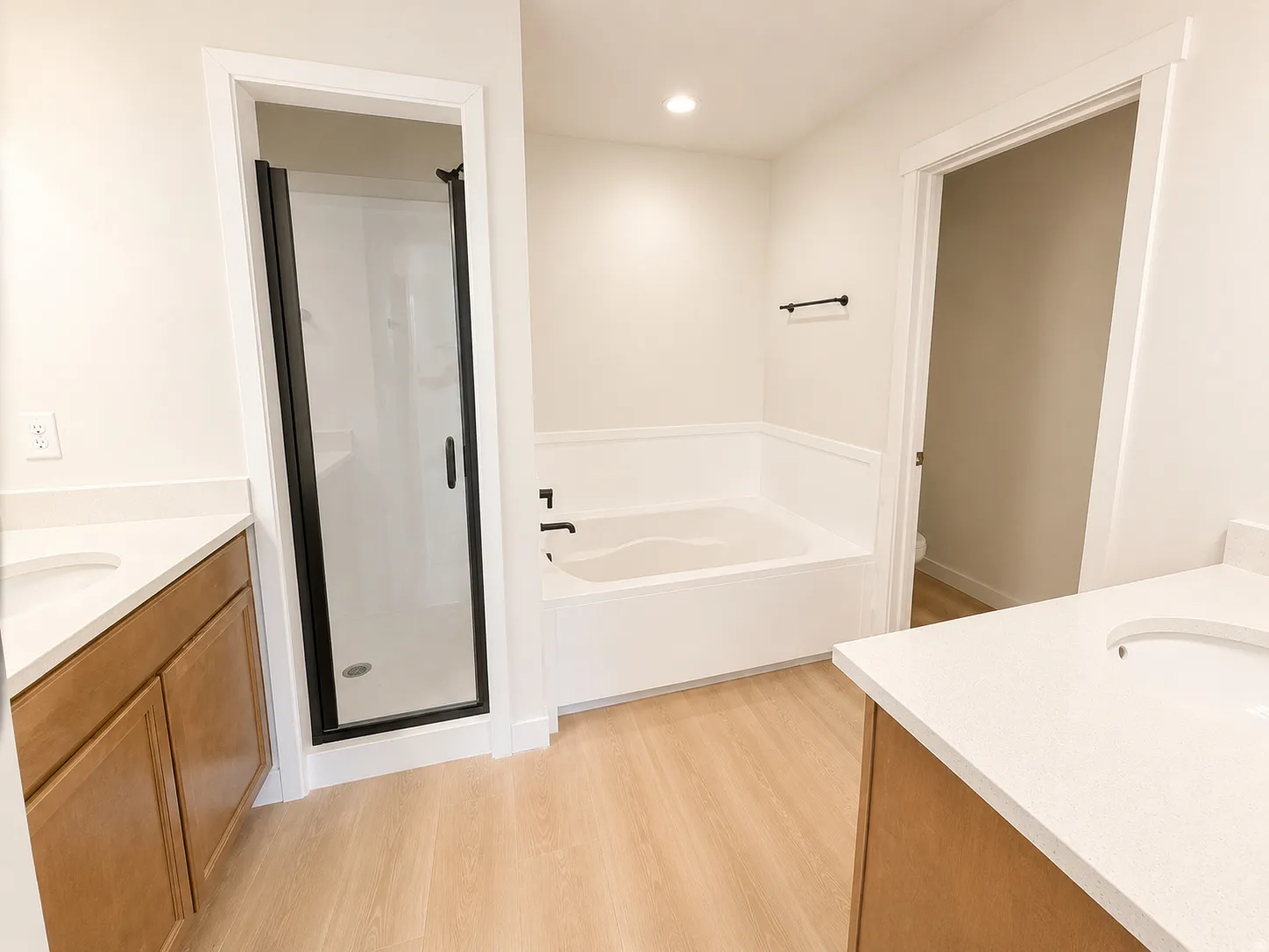 Dual vanity with integrated sinks and light wood-tone cabinetry, a built-in tub, a glass-enclosed shower with a black frame, and wood-finish flooring