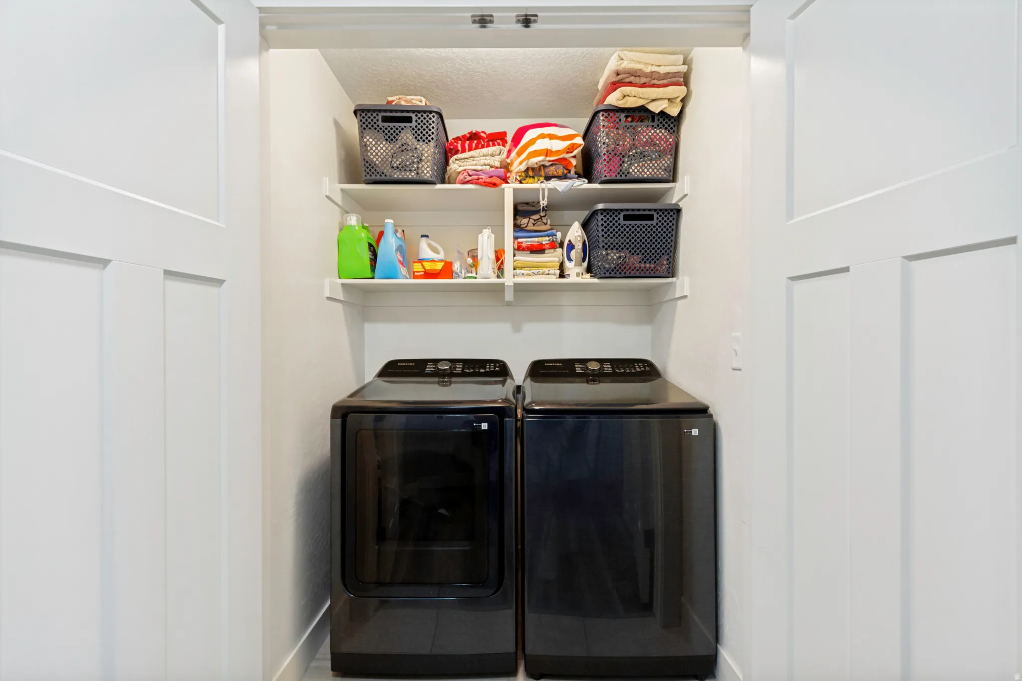 Laundry closet with a front-loading washer and dryer set, two white organizational shelves, and bright overhead lighting
