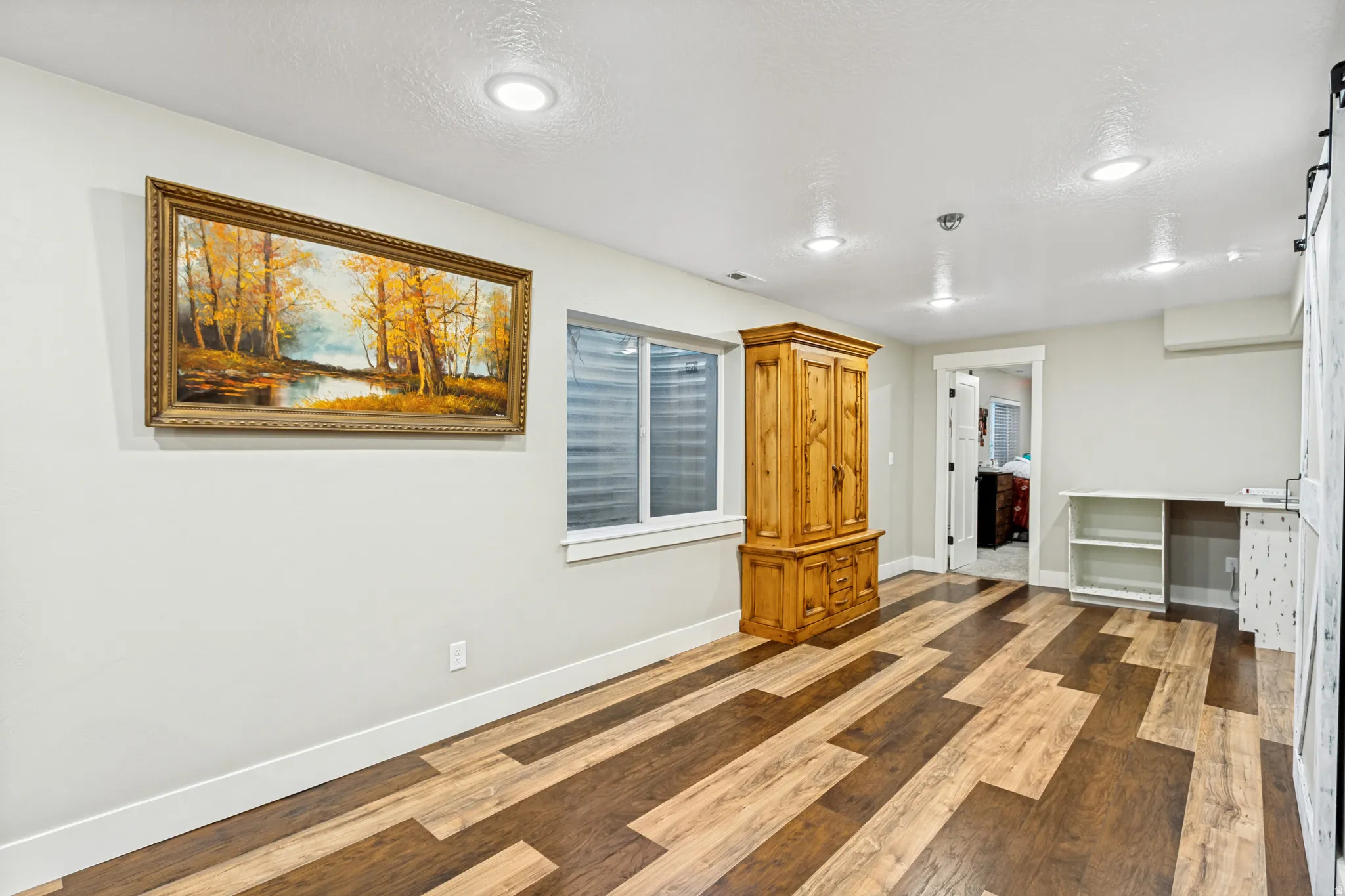 Spacious room with varied-tone wood-finish flooring, recessed lighting, a built-in desk with shelving, and a window providing natural illumination