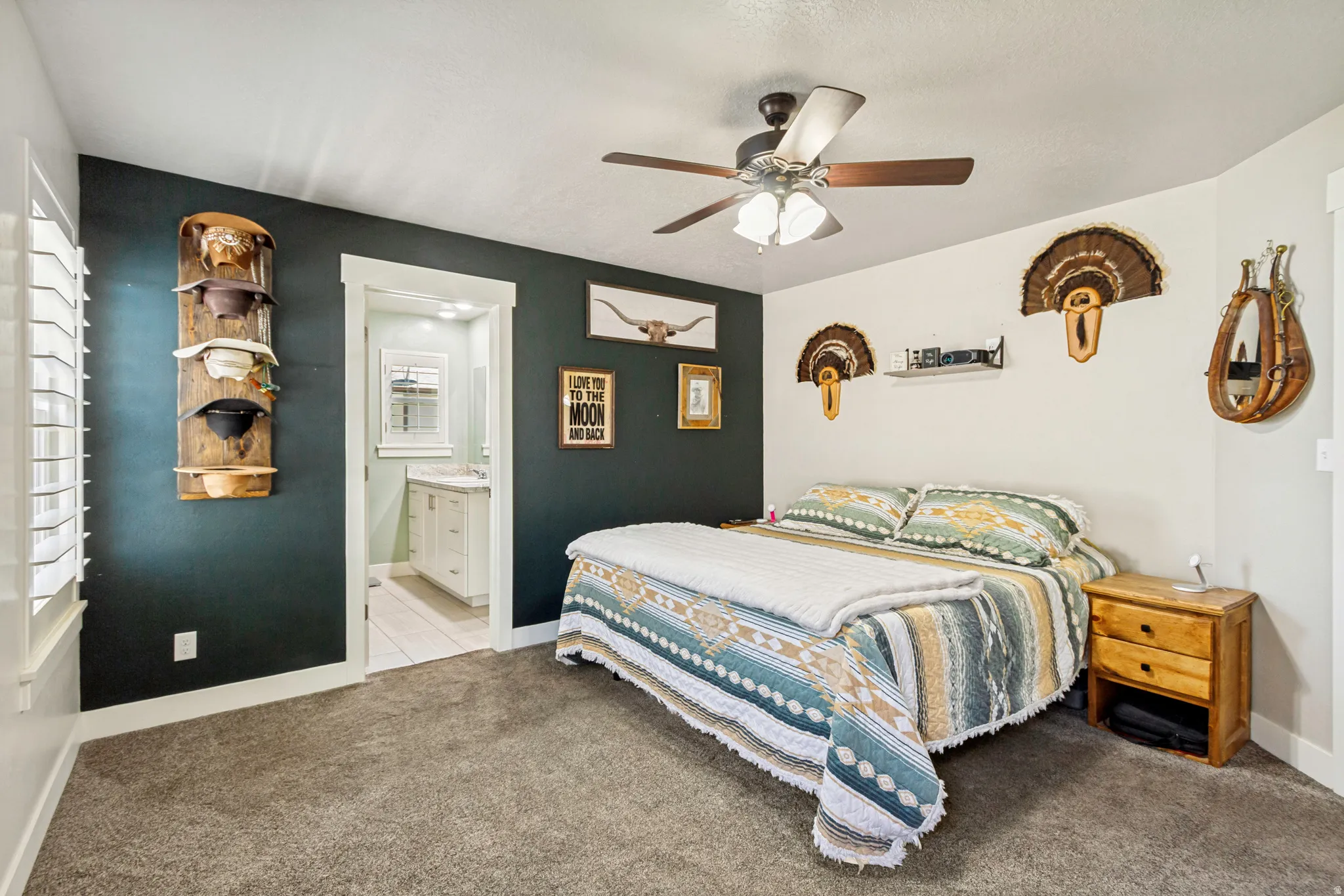 Carpeted bedroom featuring a ceiling fan with integrated lighting, a dark accent wall, white window shutters, and a private en-suite bathroom with a white vanity and mirror