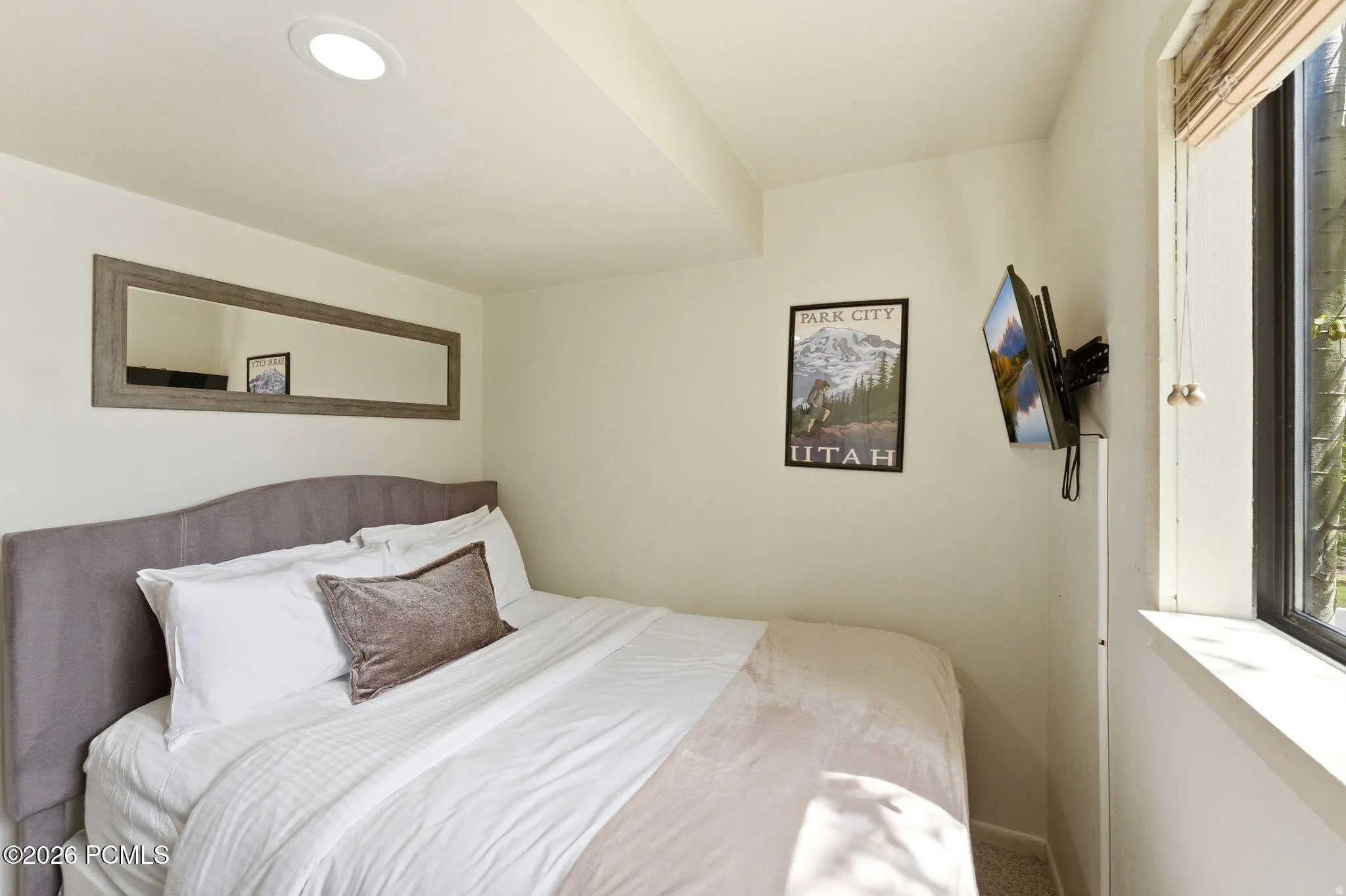 Bedroom featuring a built-in wall-mounted television, a large window with blinds, and a framed mirror