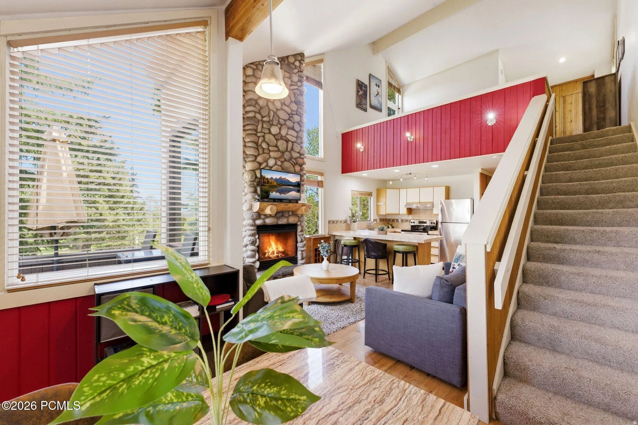 Vaulted ceiling living area featuring a prominent stone fireplace, wood-finish flooring, and expansive windows