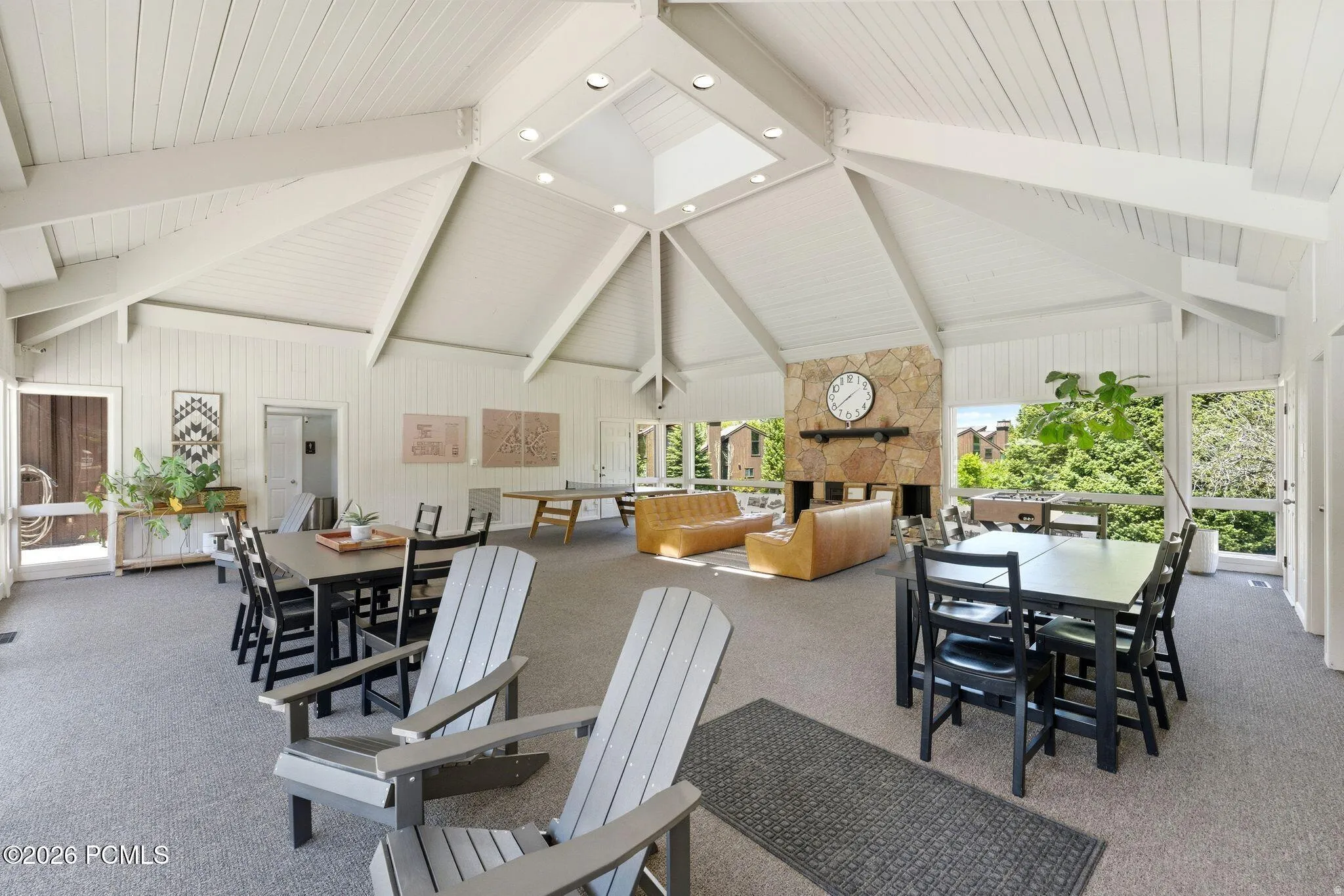 Spacious common area featuring an expansive vaulted ceiling with exposed beams, a central skylight, a stone-faced fireplace, and large windows