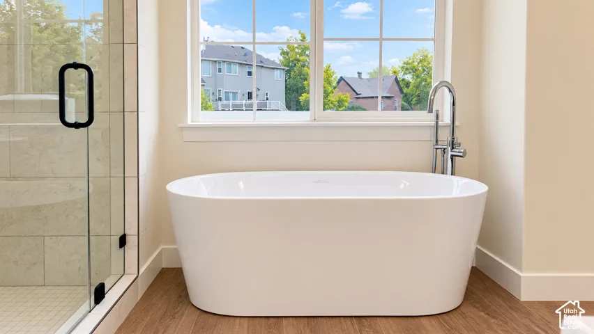 Freestanding white soaking tub with polished chrome floor-mounted faucet, wood-finish flooring, and glass-enclosed shower with tiled surround