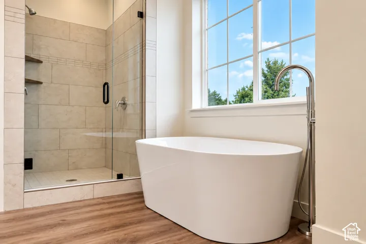 Freestanding soaking tub with a floor-mounted faucet, glass-enclosed shower with tile surround, built-in wooden shelving, and wood-finish flooring