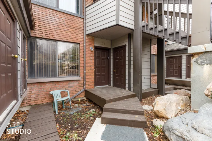 Exterior entrance featuring brick and horizontal siding, multiple brown paneled entry doors, an upper-level balcony with wood railing, and a ground-level wood walkway with steps