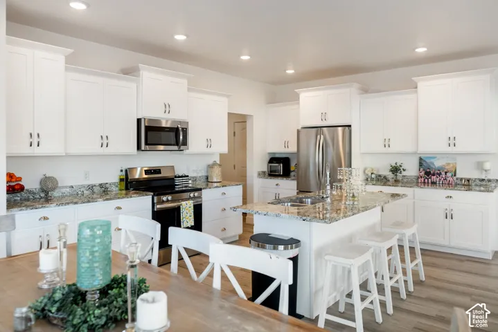 Bright kitchen featuring white shaker cabinetry, granite countertops, stainless steel appliances, a center island with a sink and bar seating, and wood-finish flooring