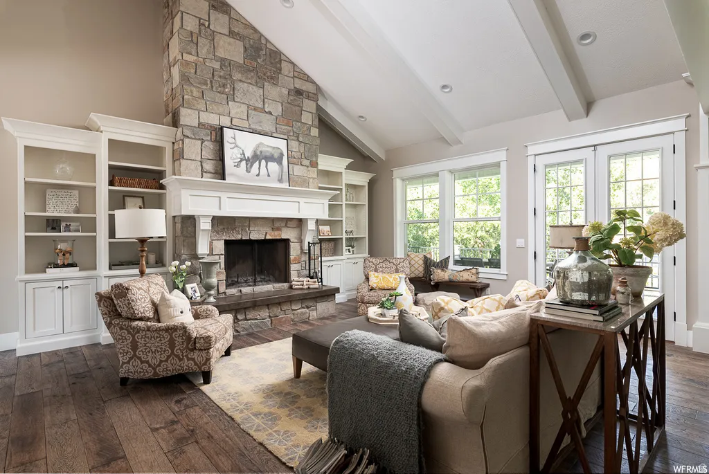 Vaulted ceiling featuring exposed beams, floor-to-ceiling stone fireplace, wood-finish flooring, custom built-in cabinetry, and multiple windows
