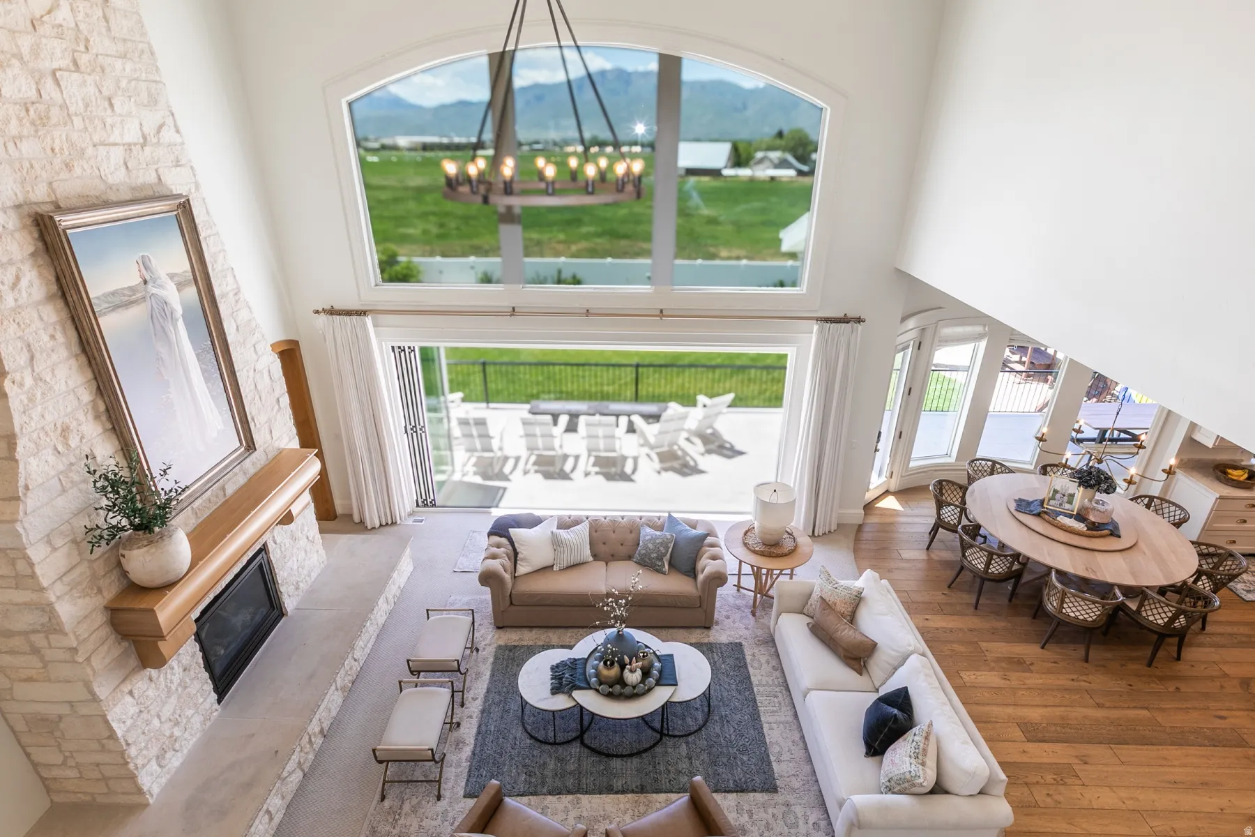 Two-story open concept living space featuring a floor-to-ceiling stone-clad fireplace with a wood mantle, expansive windows, large sliding glass doors, hardwood flooring, and a contemporary chandelier