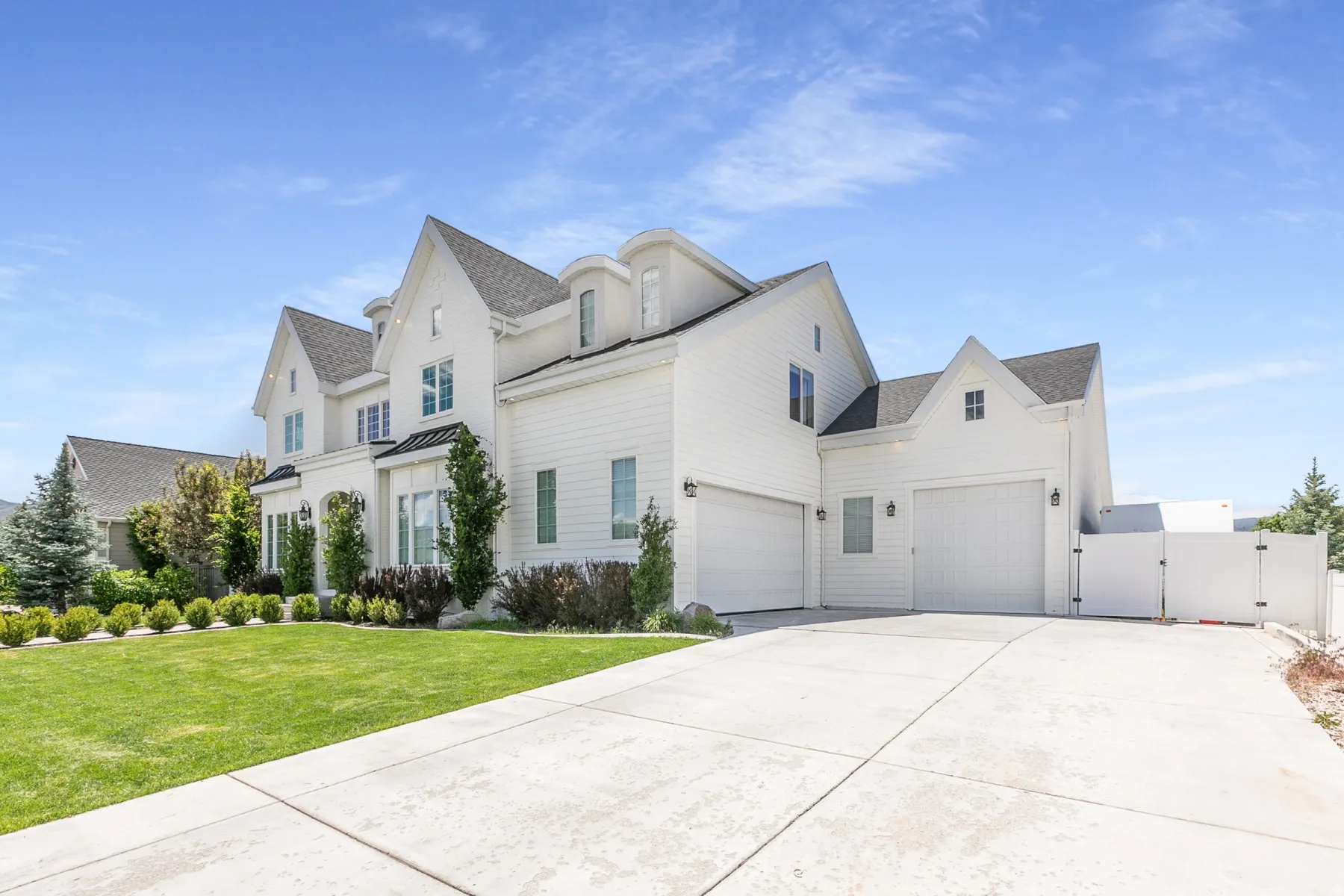 White exterior residence featuring multiple gables, a multi-car garage, manicured front lawn, mature landscaping, and an extended driveway