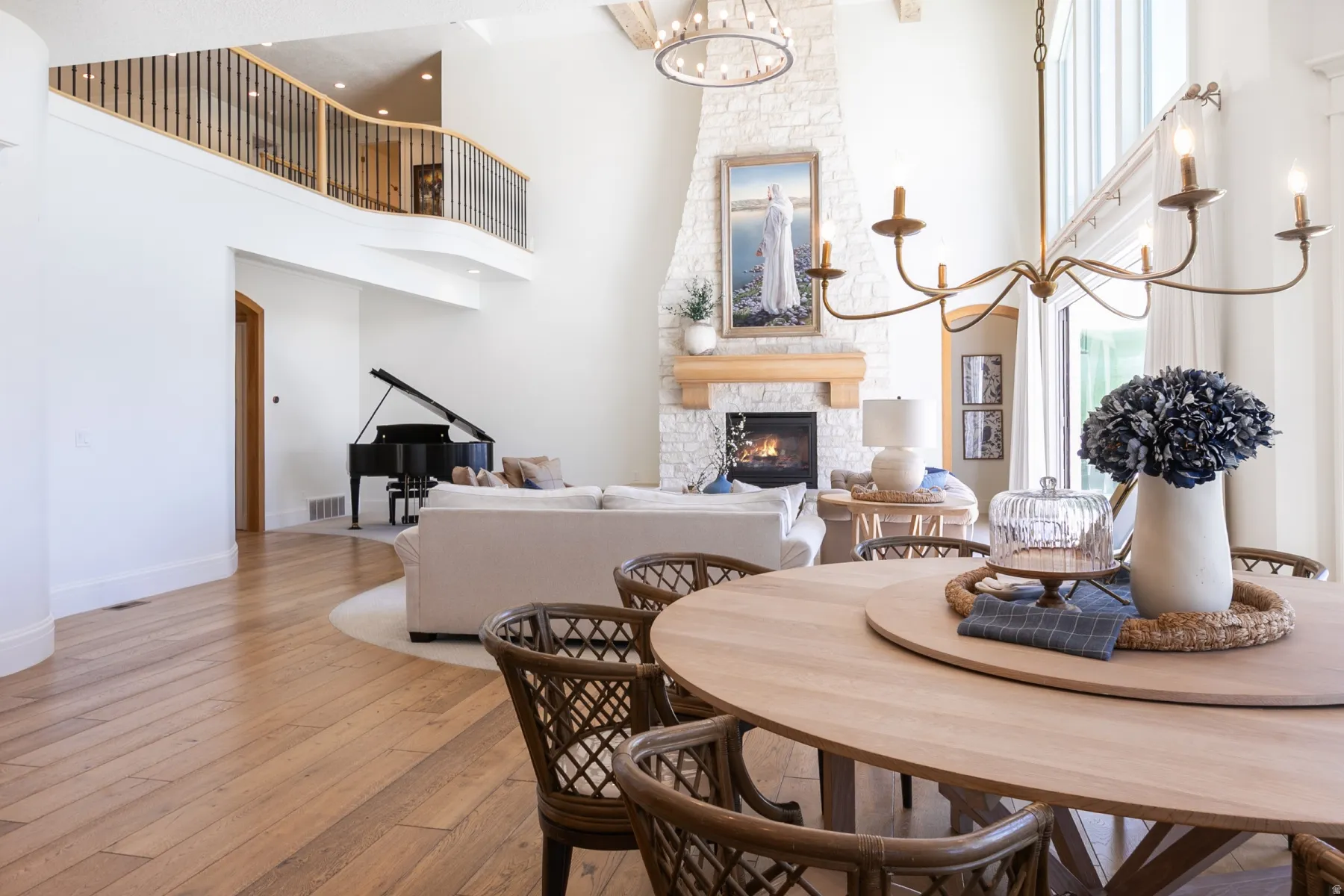 Open-concept living area with wood-finish flooring, a two-story stone fireplace, and a second-story balcony featuring a wood and metal railing