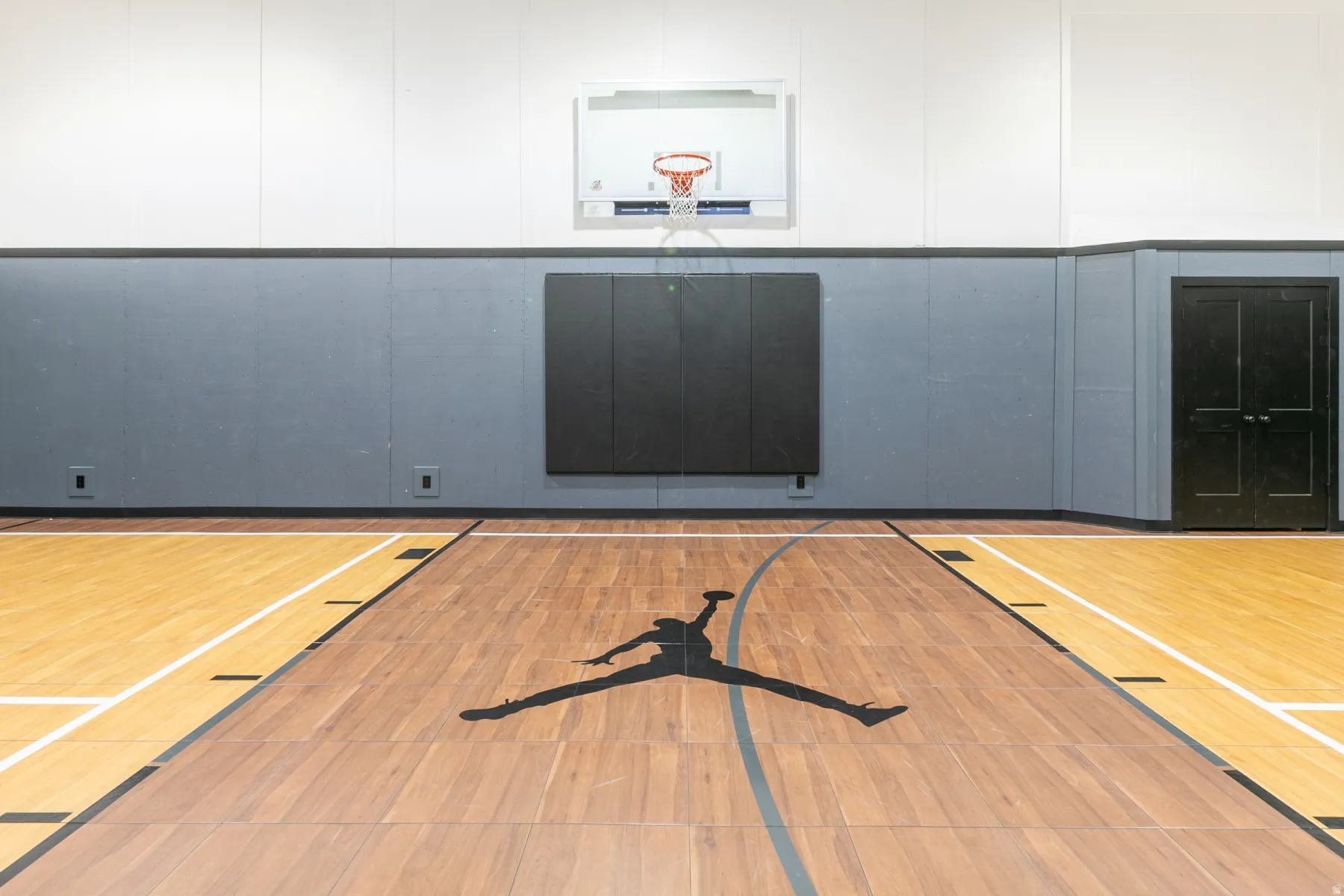 Indoor sports court featuring hardwood flooring with court markings, a wall-mounted basketball hoop, black padded wall protection, and paneled walls
