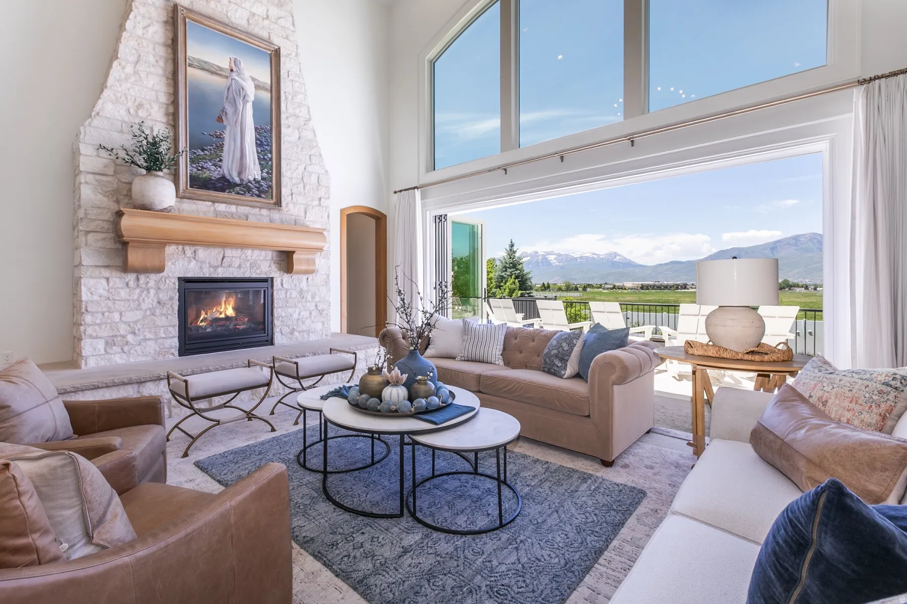 Spacious interior featuring a stone-clad fireplace with a wood mantel, floor-to-ceiling windows, and multi-panel glass doors opening to an outdoor patio with mountain scenery