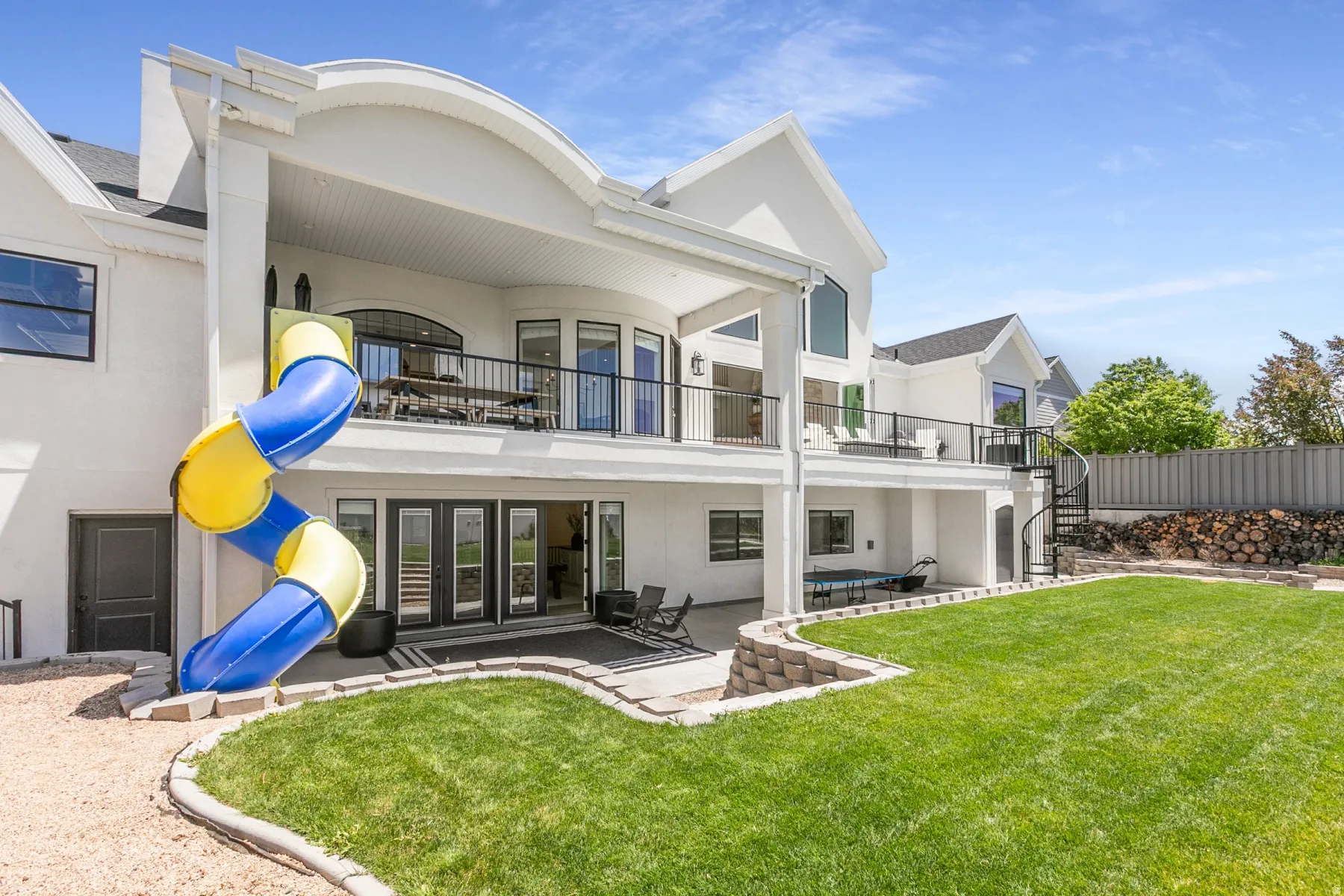 Expansive rear exterior featuring a manicured lawn, multi-level patios, a spiral staircase, an integrated slide, and a second-story balcony with dark railings