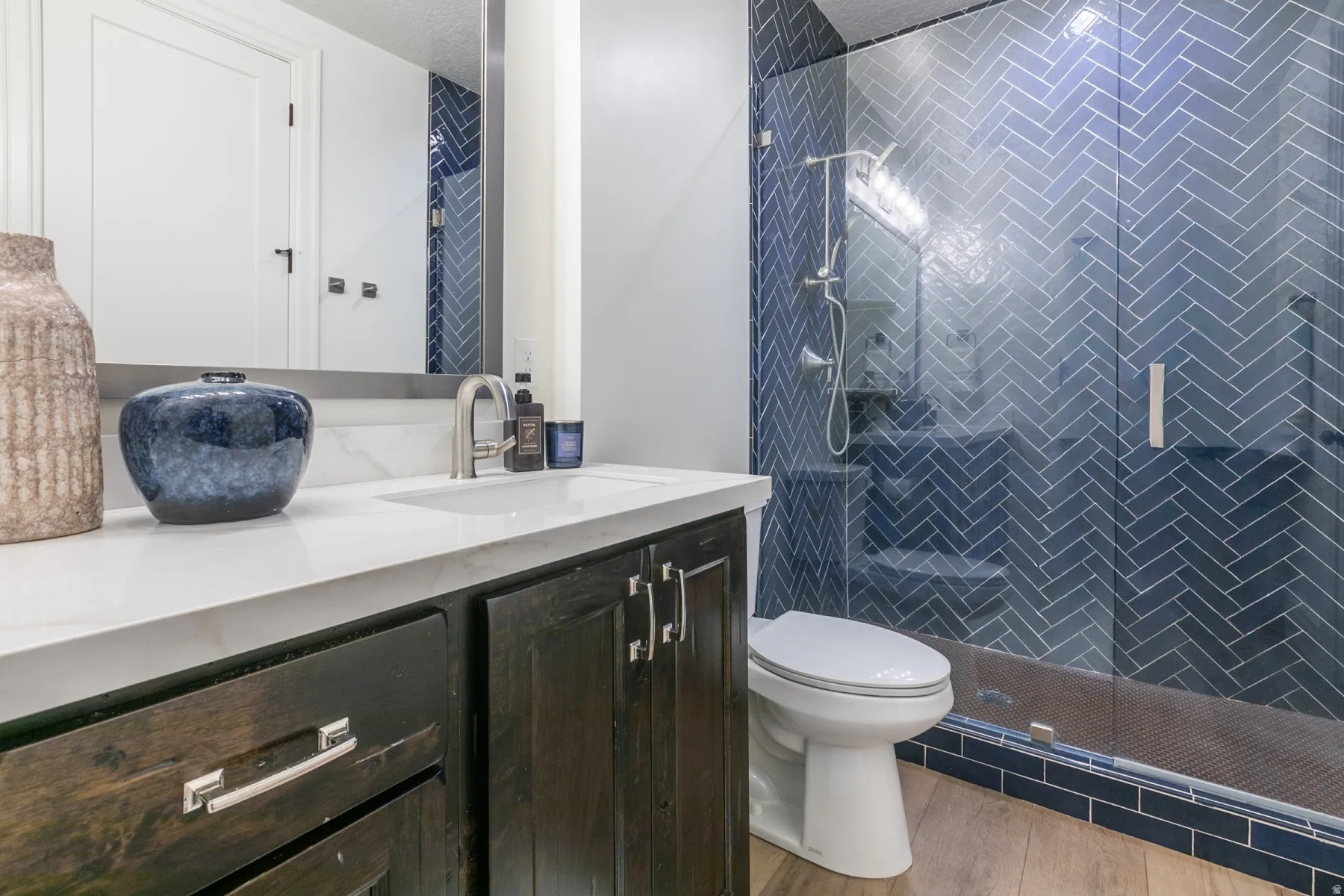 Bathroom featuring a glass-enclosed shower with blue herringbone tile, a dark wood-finish vanity, a white countertop, and a rectangular undermount sink