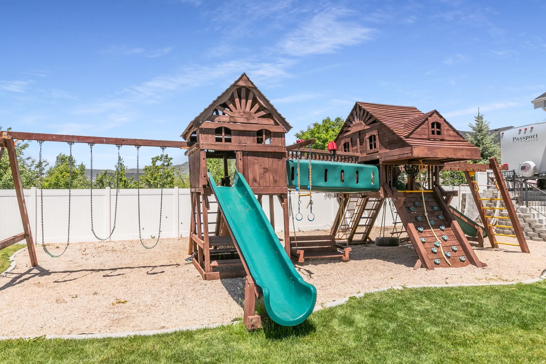 Expansive backyard featuring a multi-level wooden play structure with two slides, a rock climbing wall, and a swing set