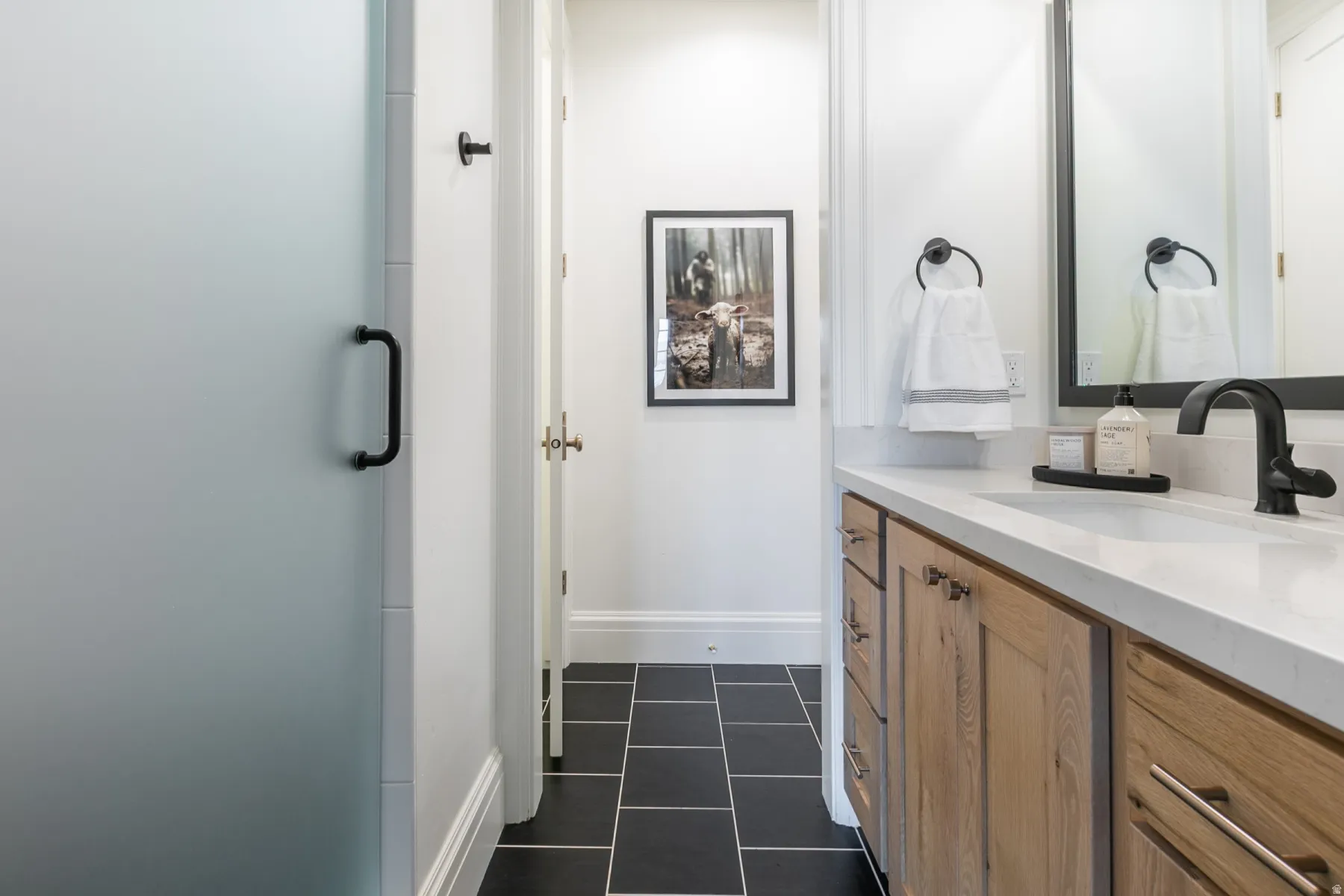 Frosted glass shower enclosure with matte black hardware, wood-finish vanity cabinetry, white countertop, black square floor tiling, and a large wall-mounted mirror