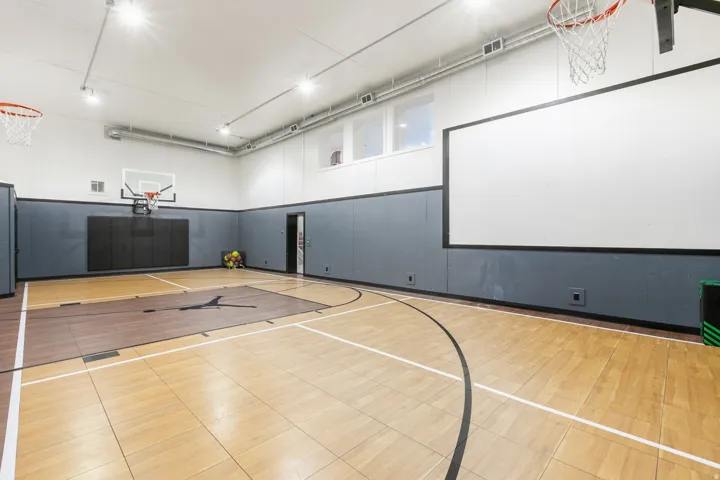Indoor basketball court featuring hardwood flooring, multiple hoops, a large projection screen, padded wall panels, and overhead lighting