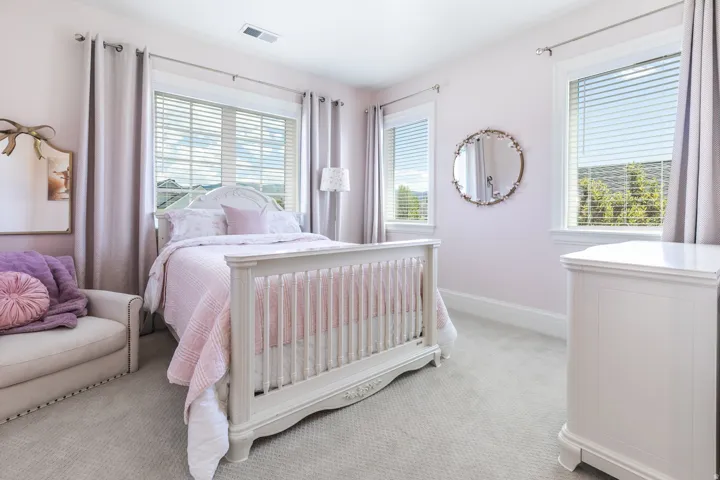 Bright bedroom featuring two large windows with blinds, light wall-to-wall carpeting, and a built-in ventilation system