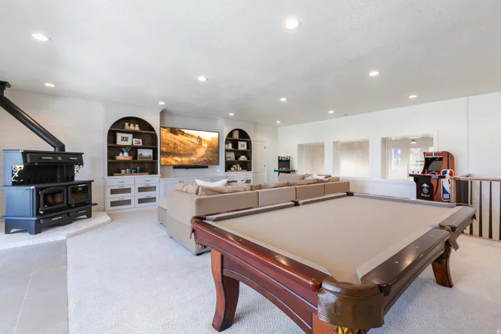 Spacious entertainment room featuring a built-in media center, recessed lighting, a decorative wood stove on a tiled hearth, and an elegant billiards table