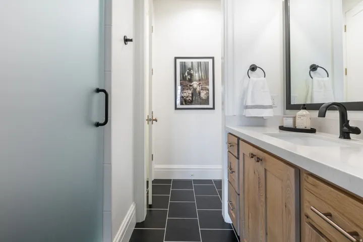 Frosted glass shower enclosure with matte black hardware, wood-finish vanity cabinetry, white countertop, black square floor tiling, and a large wall-mounted mirror
