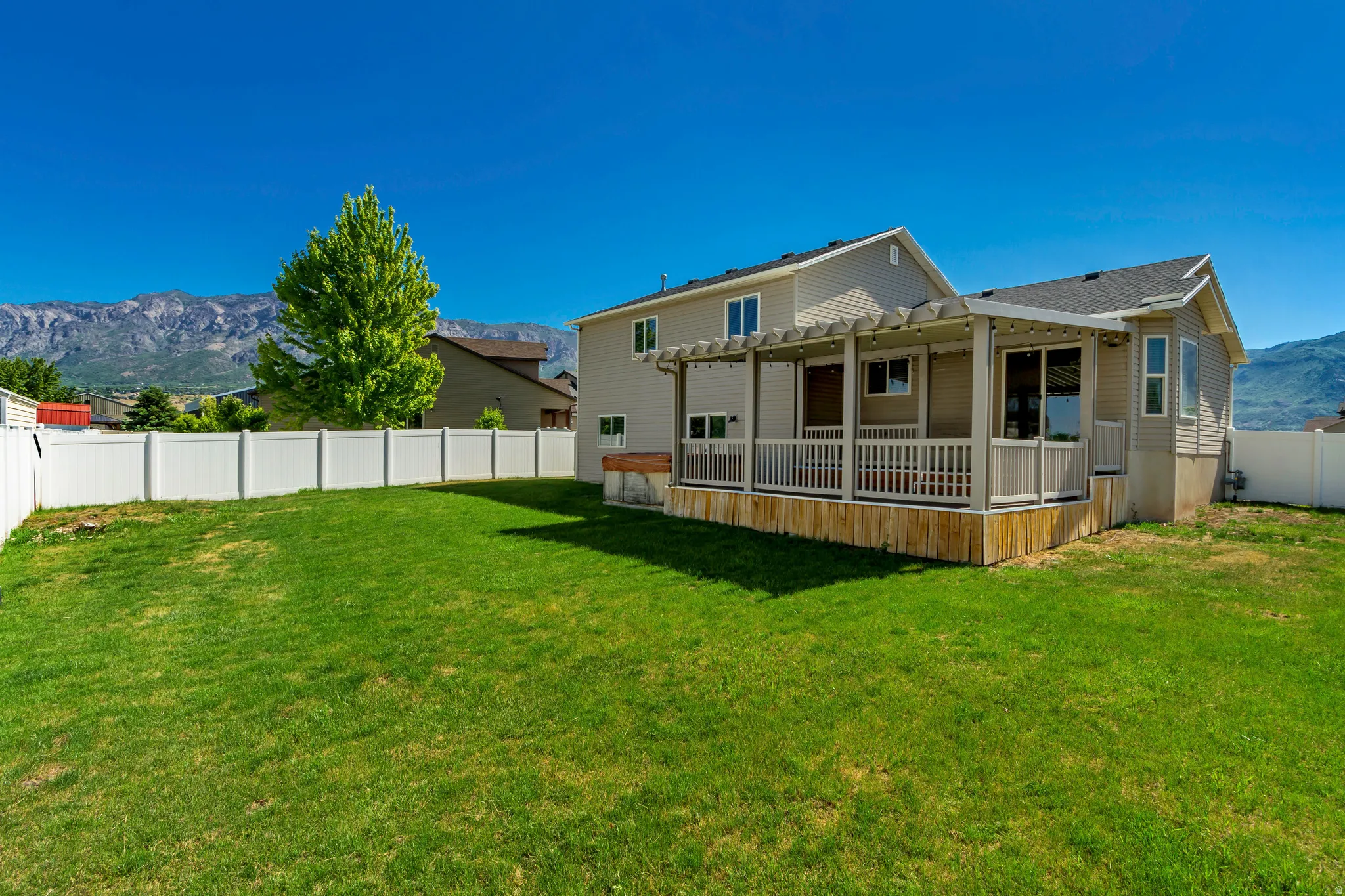 Spacious backyard featuring a manicured lawn, white vinyl fencing, a covered patio with a pergola, and mountain scenery in the distance