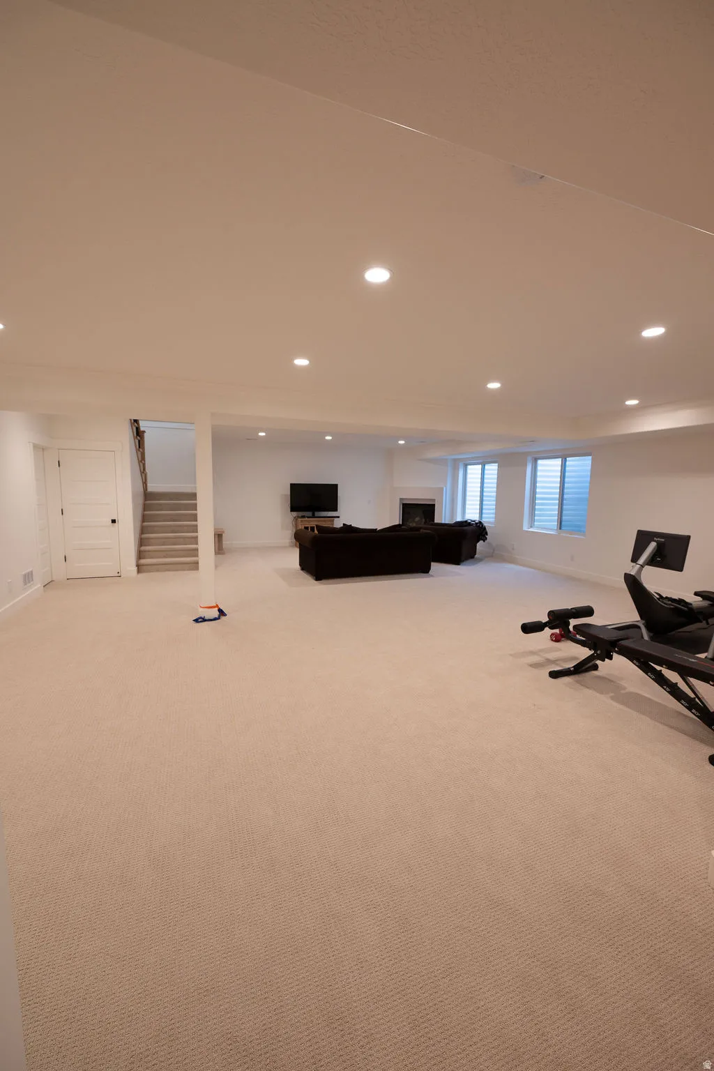 Spacious lower-level living area with recessed lighting, light-toned carpeting, white walls, and a visible staircase with wooden treads