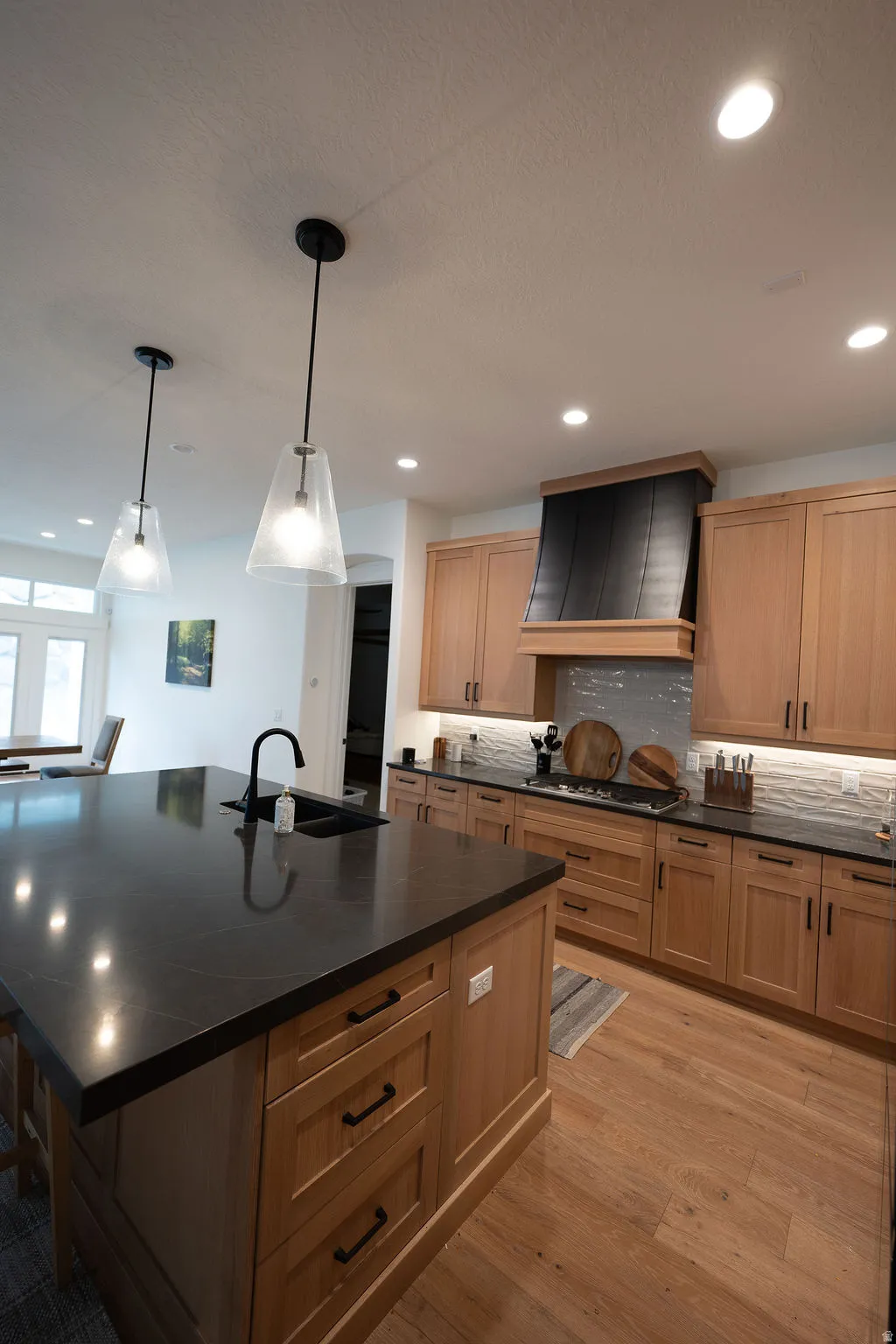 Kitchen featuring a large island with a black sink and faucet, light wood cabinetry, a black range hood, and wood-finish flooring