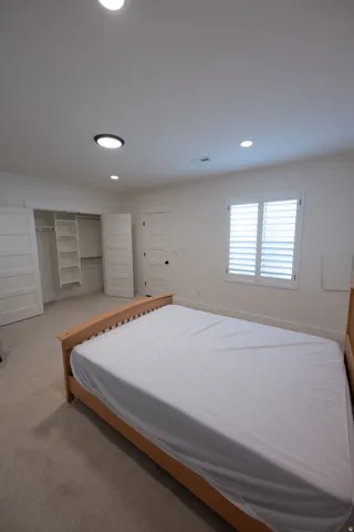 Carpeted room with a double-door closet featuring built-in shelving, recessed lighting, and a window with white plantation shutters