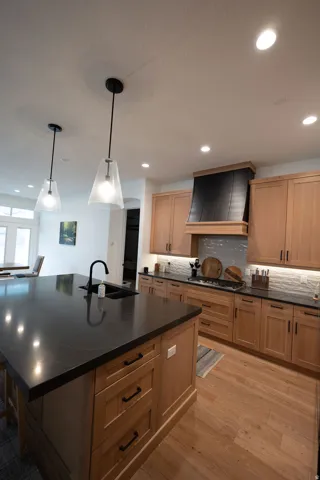 Kitchen featuring a large island with a black sink and faucet, light wood cabinetry, a black range hood, and wood-finish flooring
