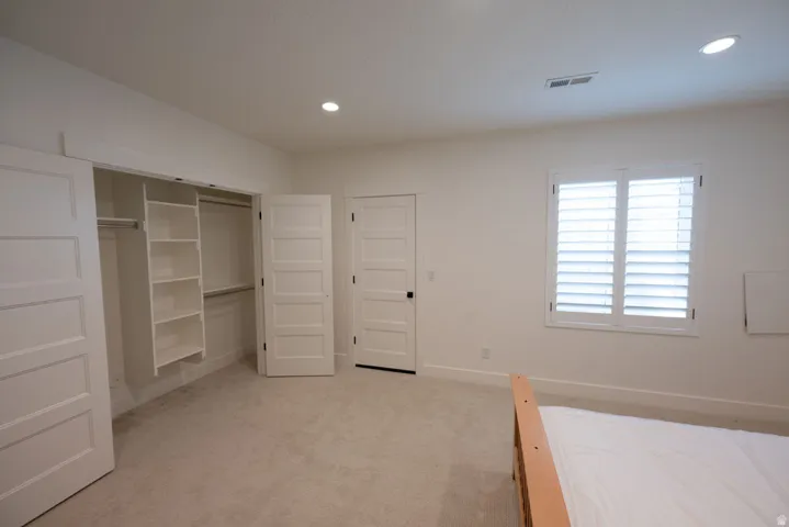Carpeted room featuring a built-in closet system with shelving and hanging rods, white louvered window shutters, recessed lighting, and a five-panel interior door