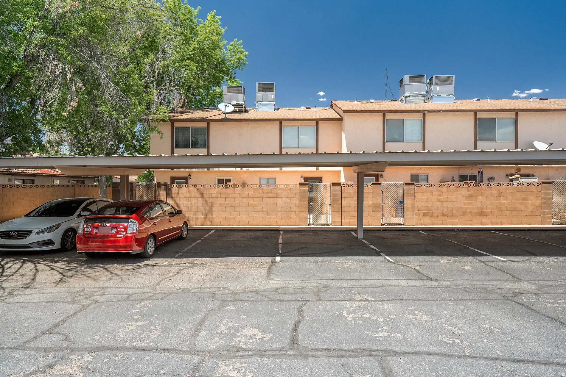 Covered parking spaces in an asphalt lot, situated in front of a multi-unit building featuring tan stucco siding and horizontal windows