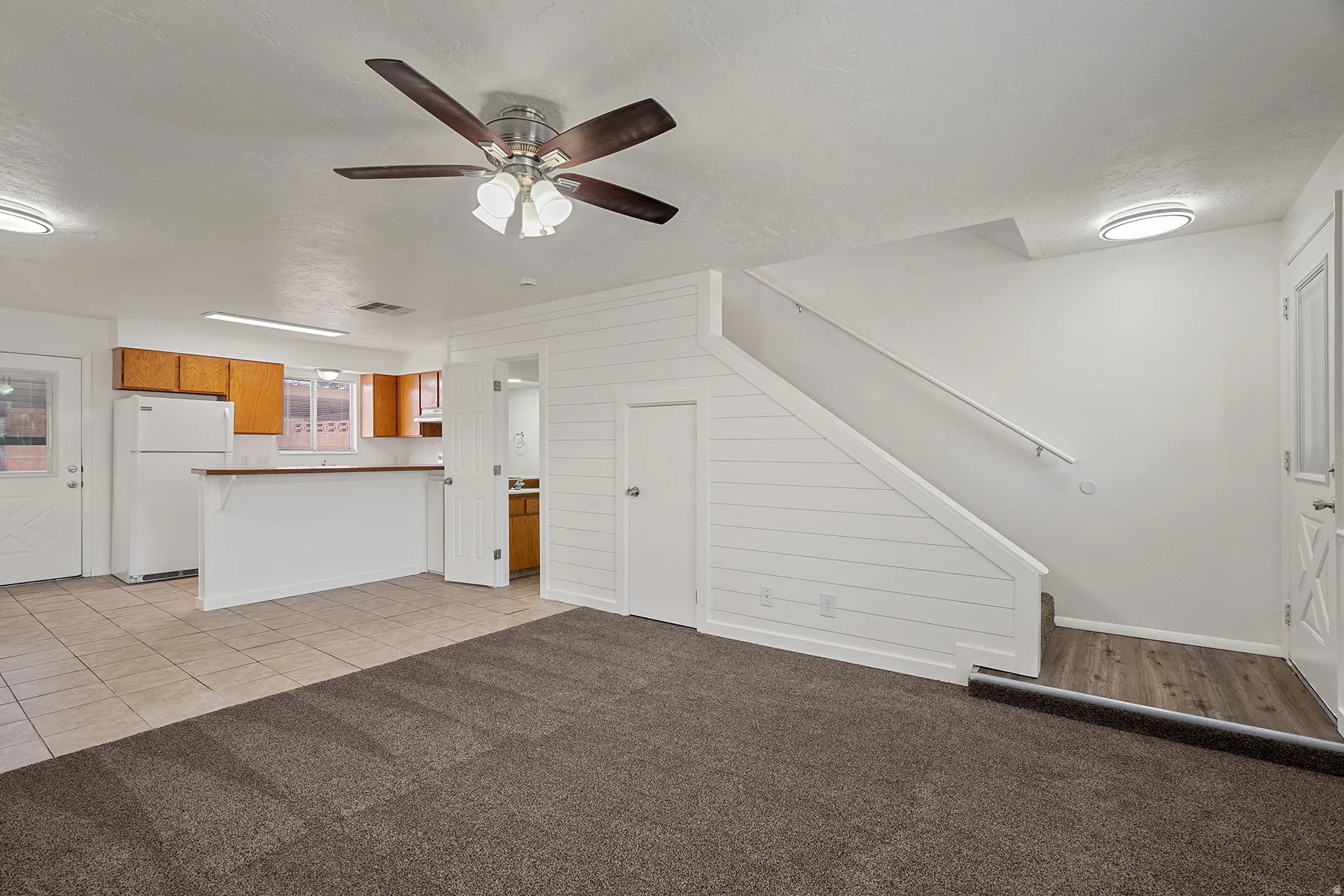 Open-concept living space featuring a ceiling fan, plush carpeting, and a kitchen area with light wood cabinetry and a breakfast bar