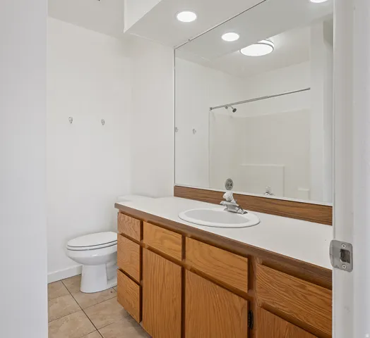 Bathroom featuring a wood-finish vanity with a single basin sink and white countertop, a toilet, a full-width wall mirror, and tile flooring