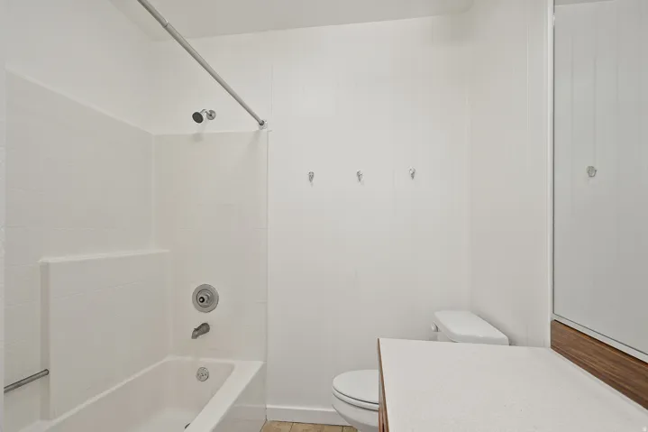 Bathroom featuring a white tub/shower combination with a textured surround, a white toilet, and a vanity with a white countertop and wood-finish base