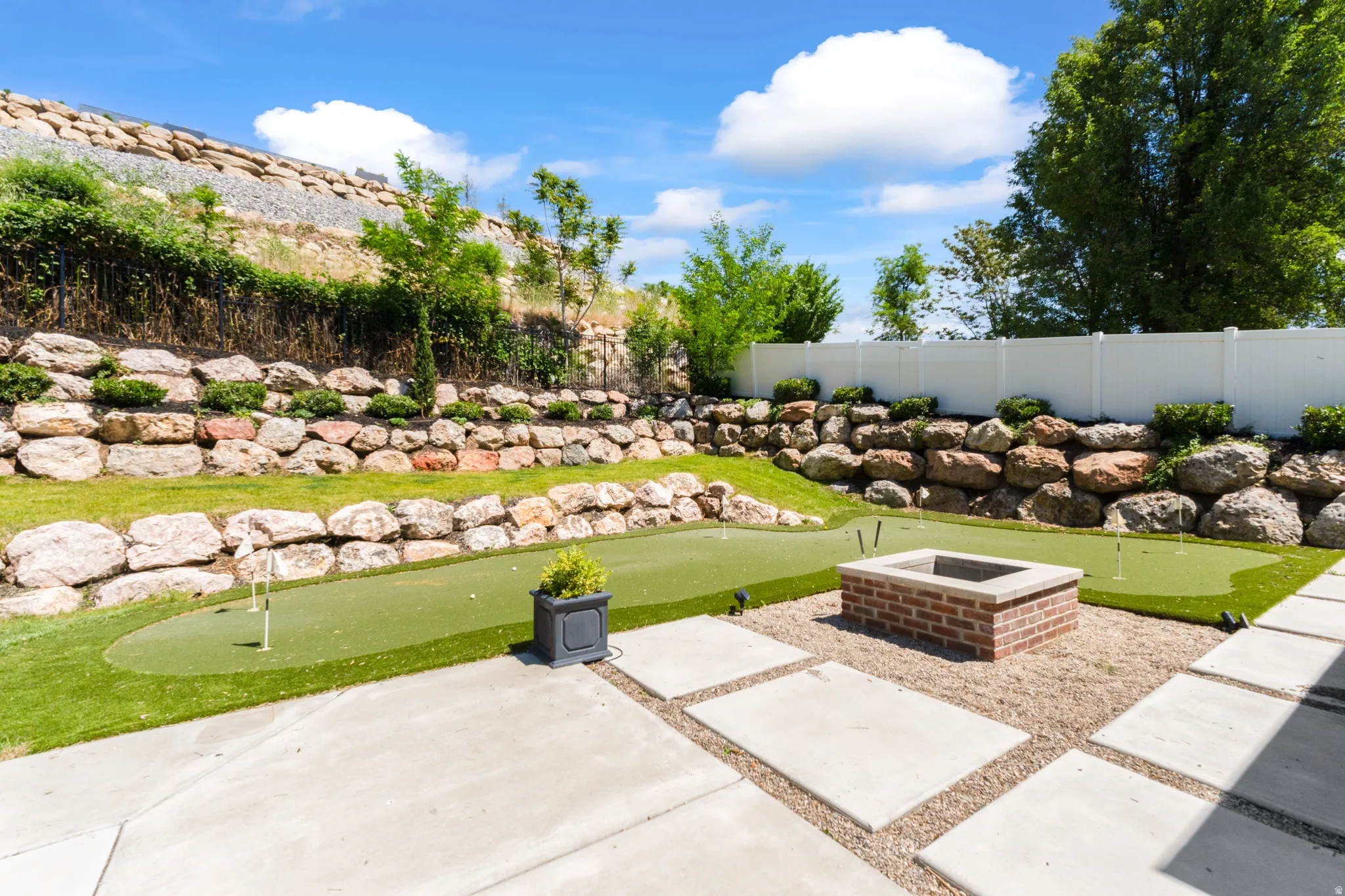 Backyard putting green and fire pit area featuring concrete pavers, gravel, and tiered rock retaining walls