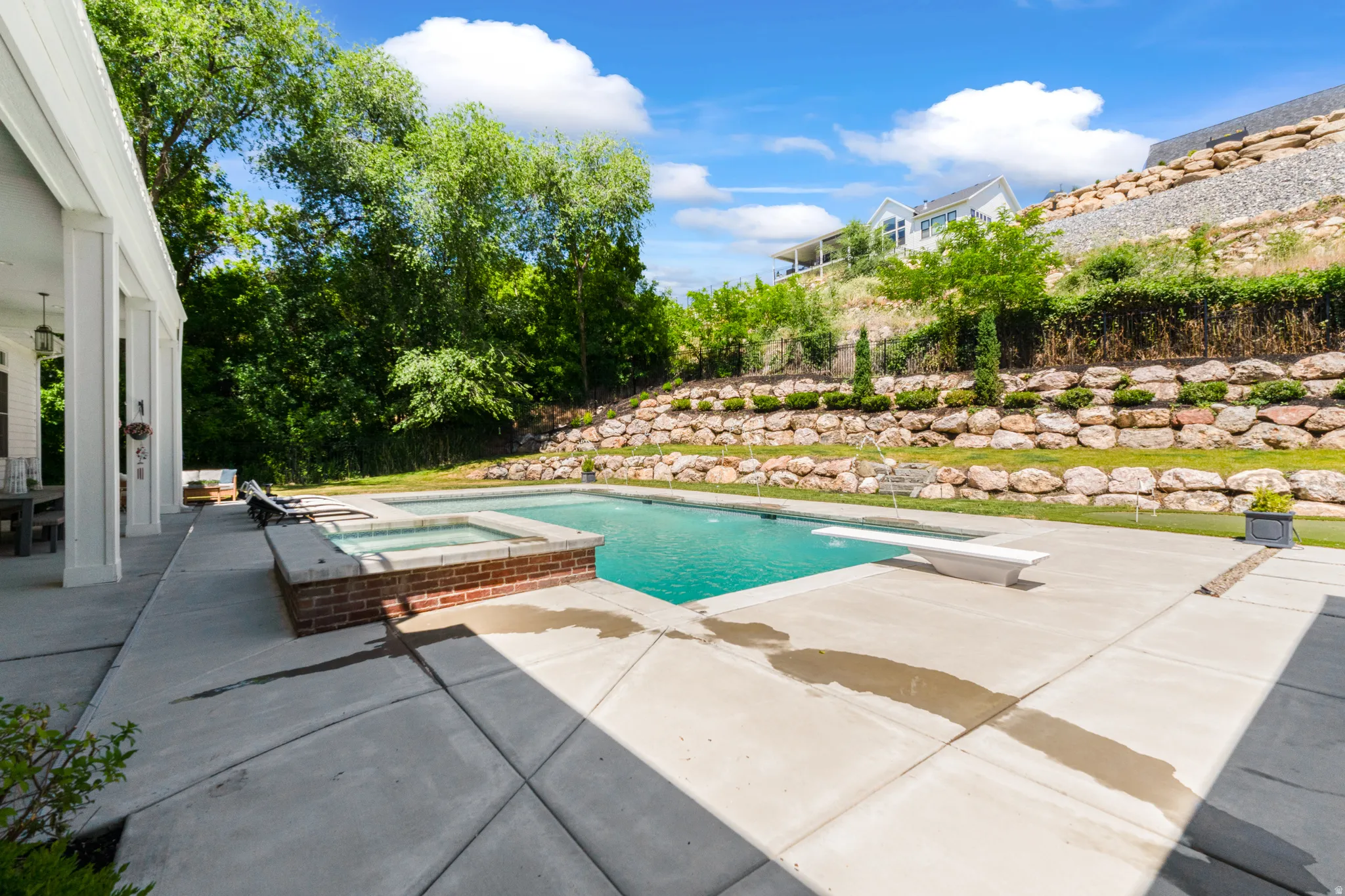 Outdoor pool area featuring a built-in spa with brick surround, concrete patio, diving board, and terraced landscaping with rock retaining walls