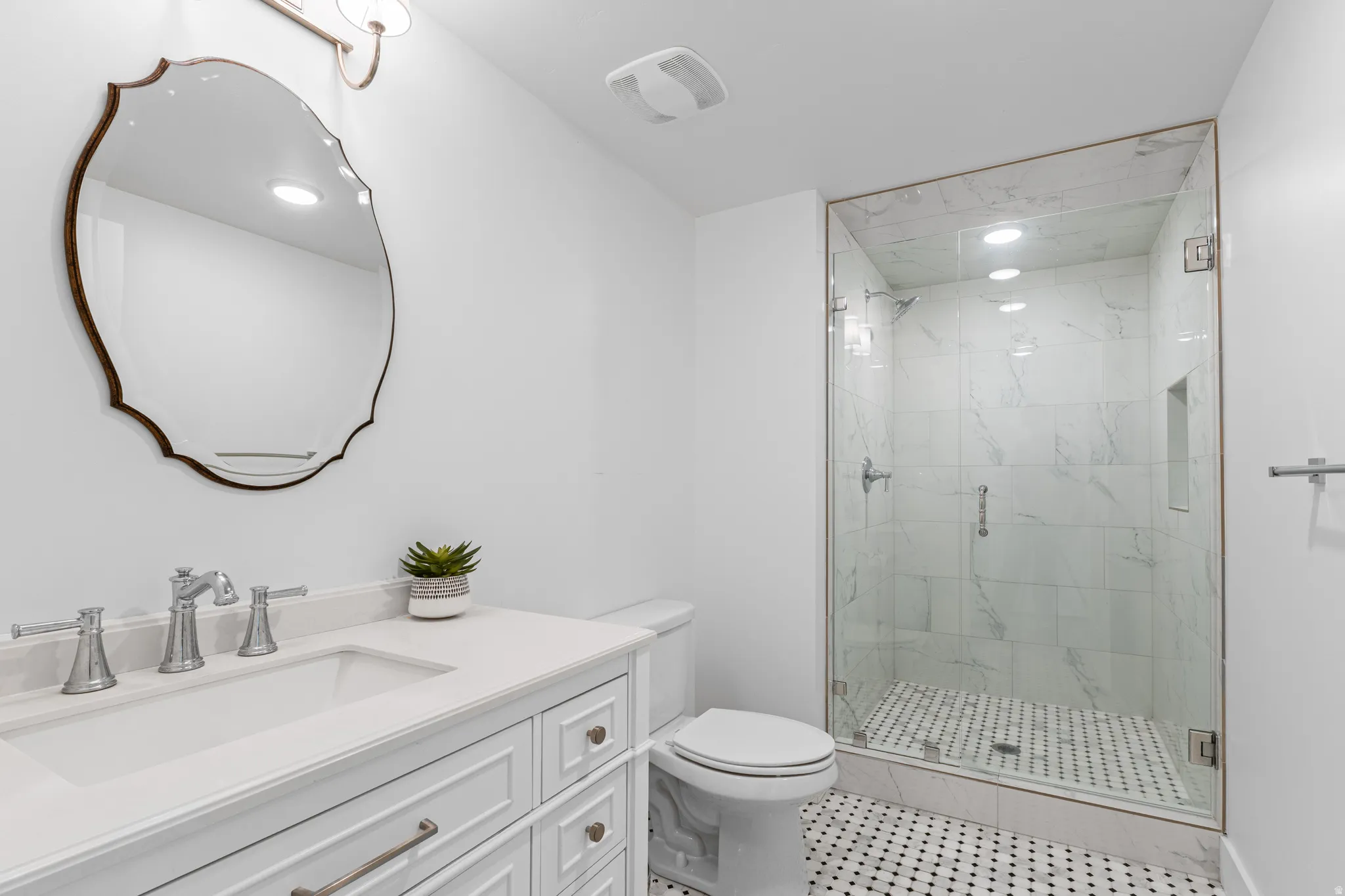 Bathroom featuring a frameless glass shower enclosure with marble-patterned tile, a white vanity with drawers, a single basin sink, and a decorative framed mirror