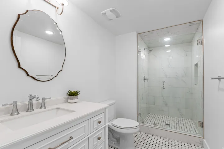 Bathroom featuring a frameless glass shower enclosure with marble-patterned tile, a white vanity with drawers, a single basin sink, and a decorative framed mirror