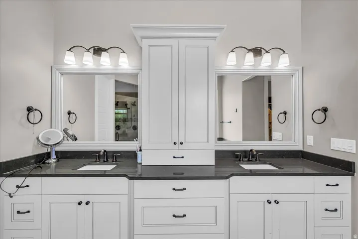 Dual vanity featuring white shaker-style cabinetry, dark countertops, undermount sinks, and wall-mounted light fixtures