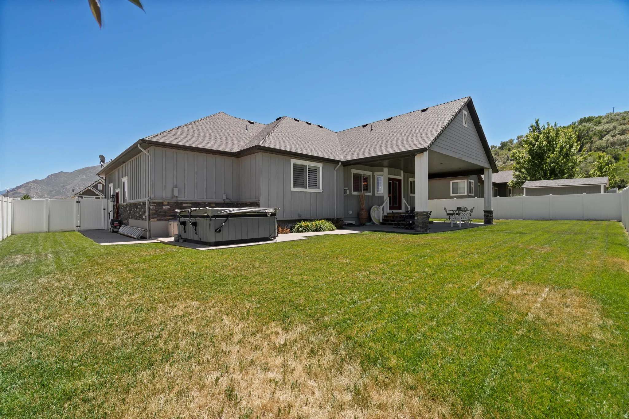 Expansive green lawn surrounds the property, featuring a covered patio with stone-wrapped pillars, a private hot tub area, and a white privacy fence