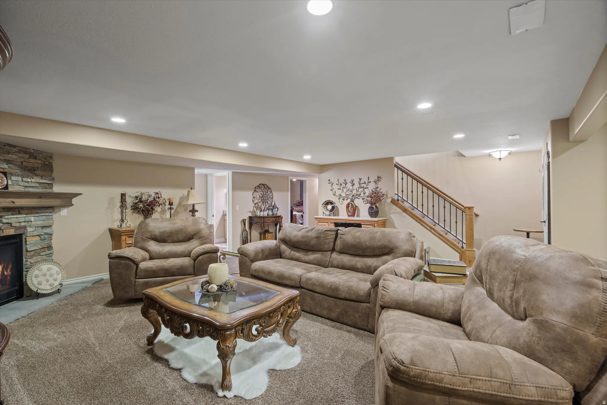 Spacious living area featuring a stacked stone fireplace with a substantial wood mantel, recessed lighting, carpet flooring, and a hardwood staircase with wrought iron balusters