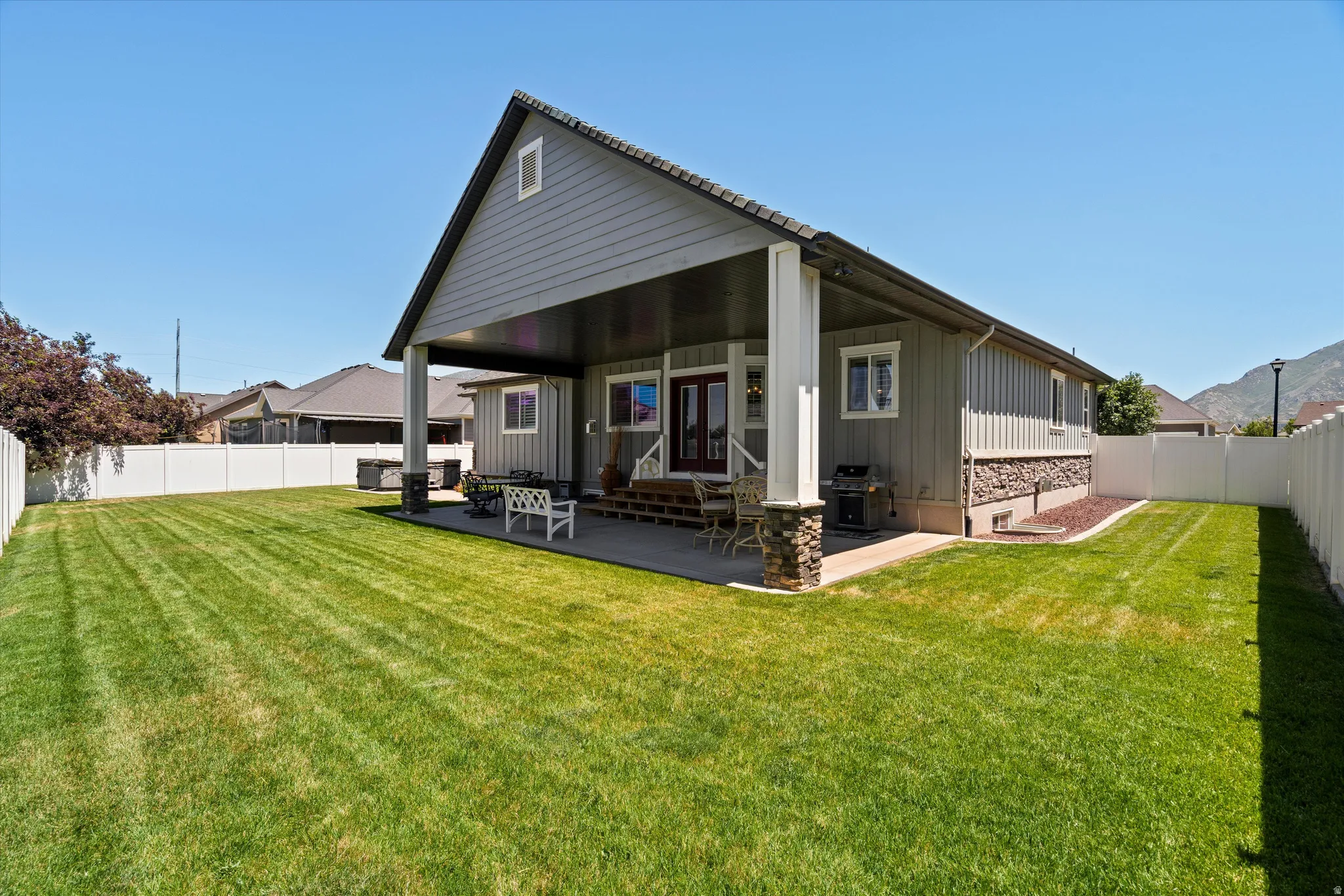 Expansive green lawn with a covered patio, featuring a vinyl privacy fence, stone accent pillars, and a mountain backdrop